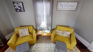 Une pièce avec deux fauteuils jaunes, une petite table d’appoint en bois sur laquelle se trouve une lampe, un panneau d’accueil encadré et un tableau floral accroché au mur.