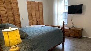 Ein Schlafzimmer mit einem Bett, einer Holzkommode, einem an der Wand befestigten Fernseher und einem Fenster mit Vorhängen.