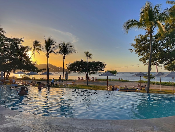 A pool area with lounge chairs and umbrellas, a view of the ocean, and palm trees.