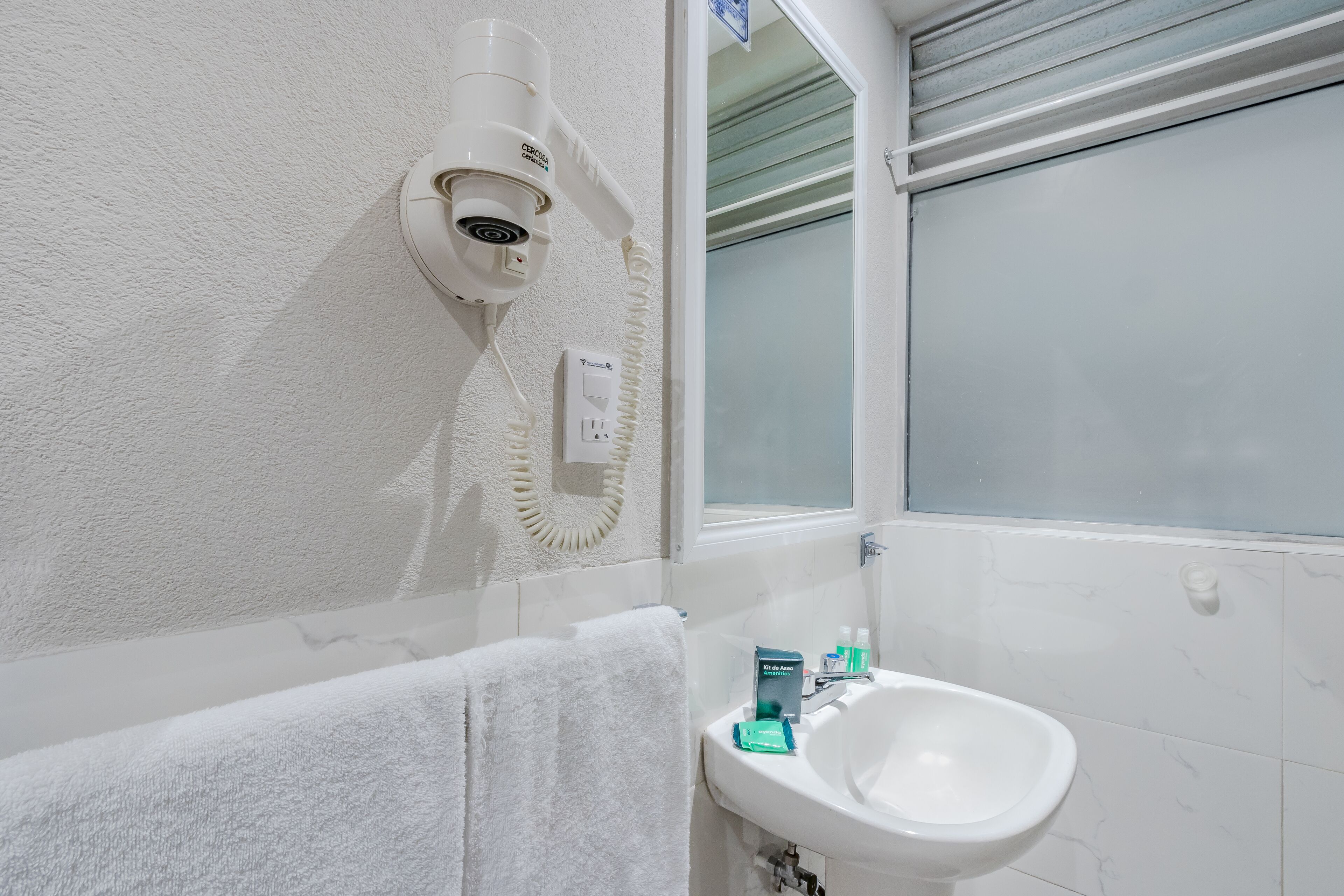 Un baño con lavamanos blanco, una toalla, una cámara de seguridad y una ventana con cristal satinado.