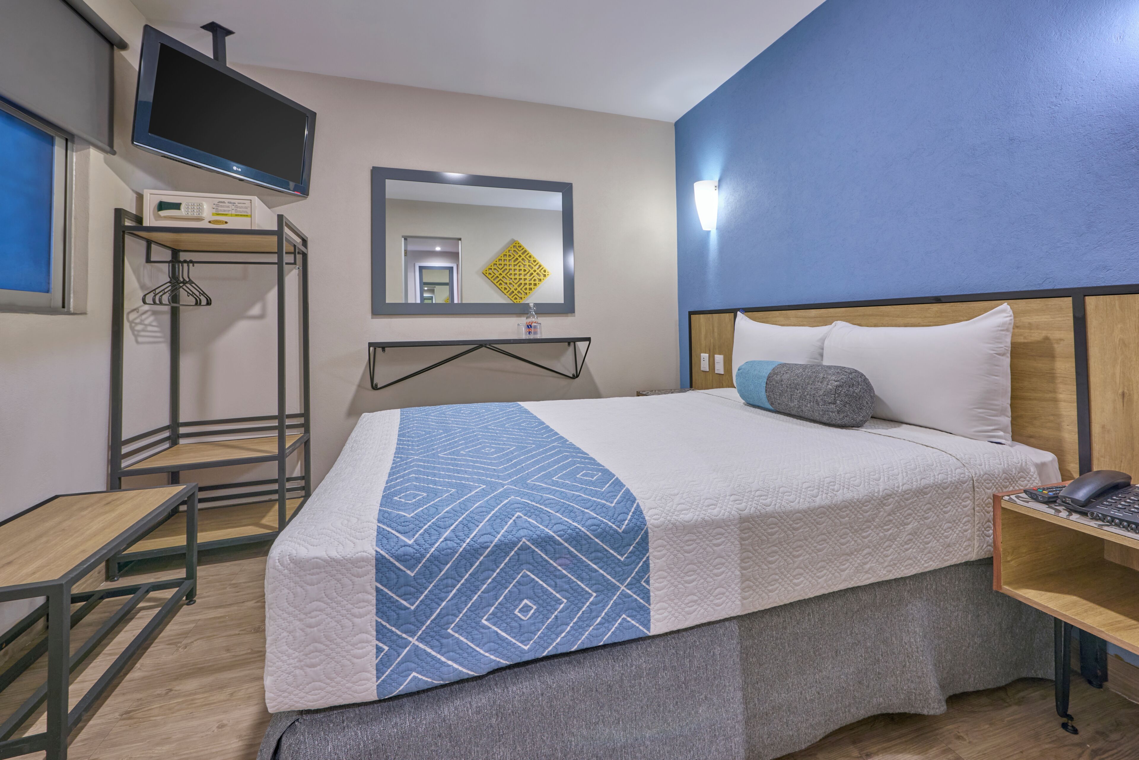 A hotel room with a bed, a TV mounted on the wall, a bedside table with a phone, and a mirror above the bed.