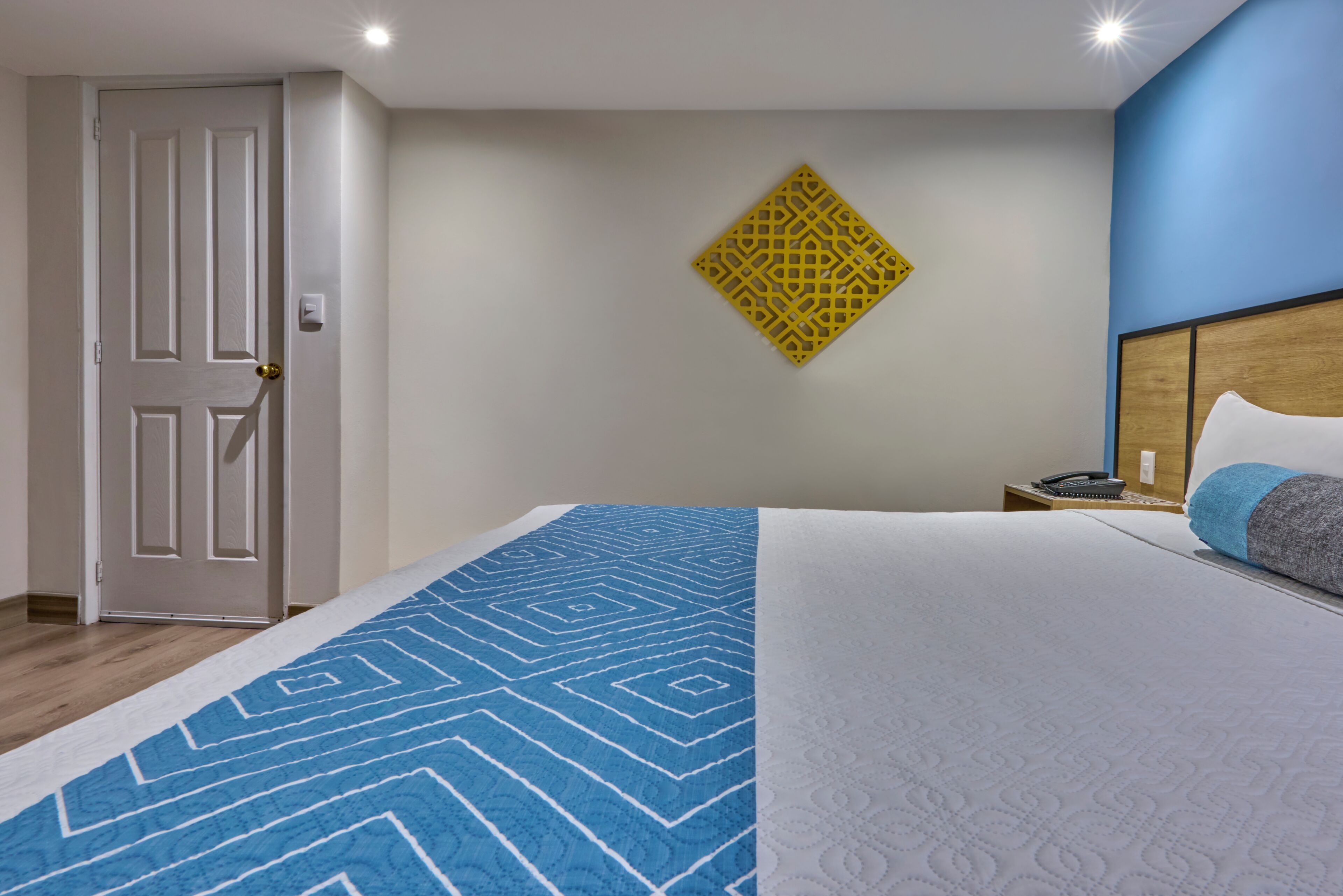 A hotel room with a bed, a door, a wall-mounted decorative item, and a bedside table with a phone and a lamp.
