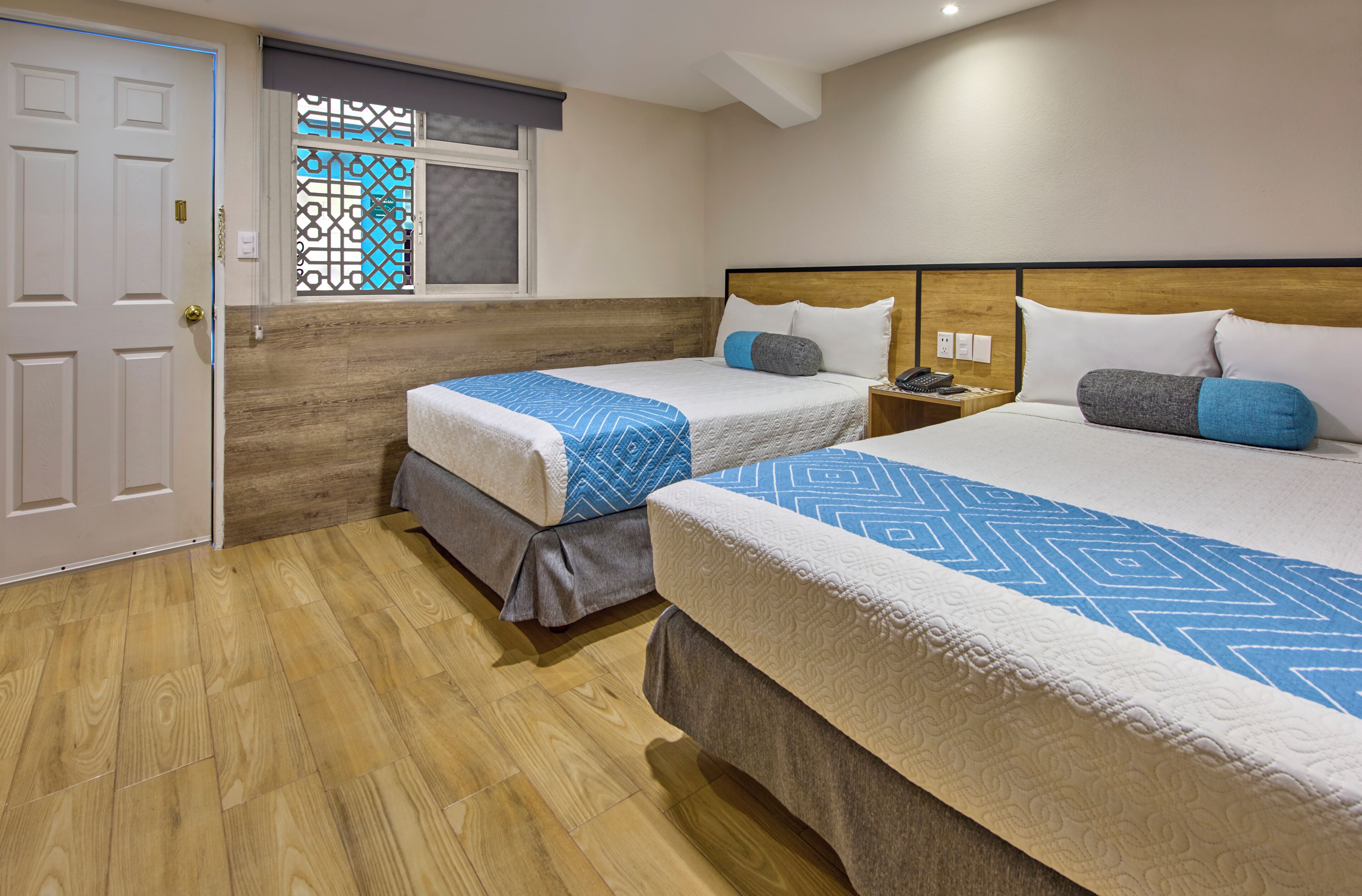 Habitación de hotel con dos camas, suelo de madera, ventana con celosía decorativa y una puerta.