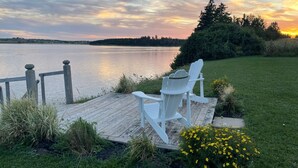 En träveranda med två vita Adirondack-stolar med utsikt över en sjö i solnedgången.