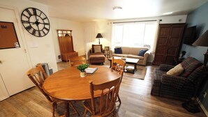 A dining area with a wooden table and chairs, a clock on the wall, and a living room with a sofa and a television. - Wooded river retreat near Mt. Bachelor and Bend (Bend)