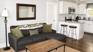 A living room with a sofa, a coffee table, a lamp, a clock, and a kitchen area with a microwave and bar stools.