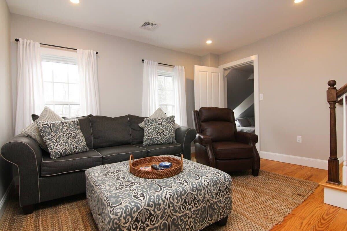 A living room with a dark grey sofa, patterned ottoman, and a brown leather chair. There is a wooden floor and a staircase in the background.