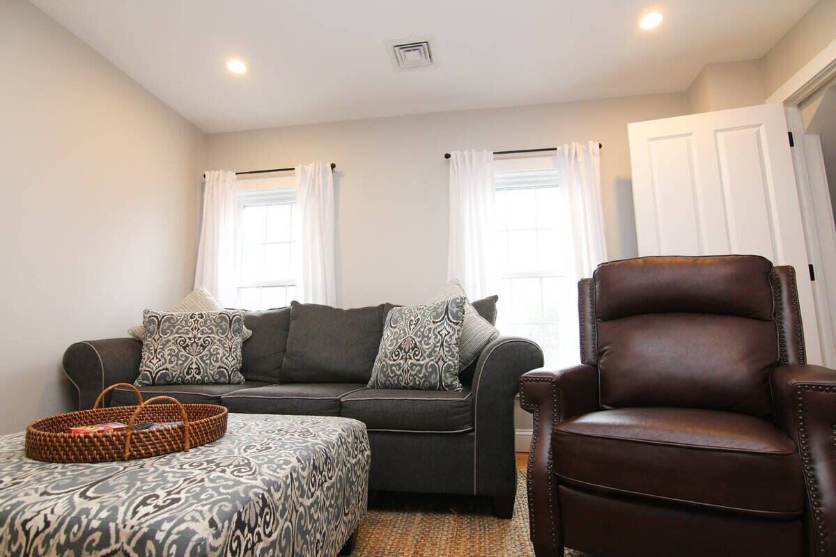 A living room with a dark brown leather armchair, a grey sectional sofa with patterned cushions, a woven basket, and a patterned ottoman.