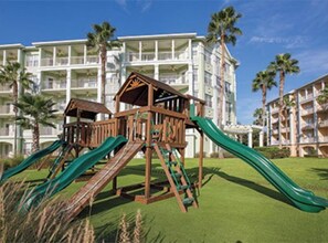 Explore the resort's playground, featuring two slides and a shelter, set against a backdrop of a multi-story hotel with balconies.
