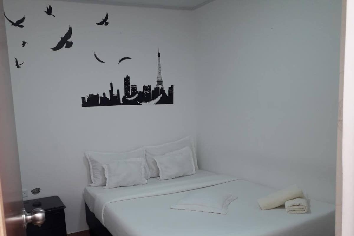 A bedroom with a bed, pillows, a nightstand, a wall decal of a city skyline, and a towel on the bed.