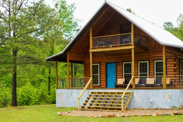 A two-story log cabin with a balcony, a blue door, and a wooden staircase leading to the entrance.