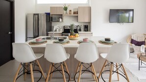 A modern kitchen with a central island, white chairs, and a flat-screen TV mounted on the wall. - Bright and stylish new condo, 5 min walk to the beach. (Jacó)