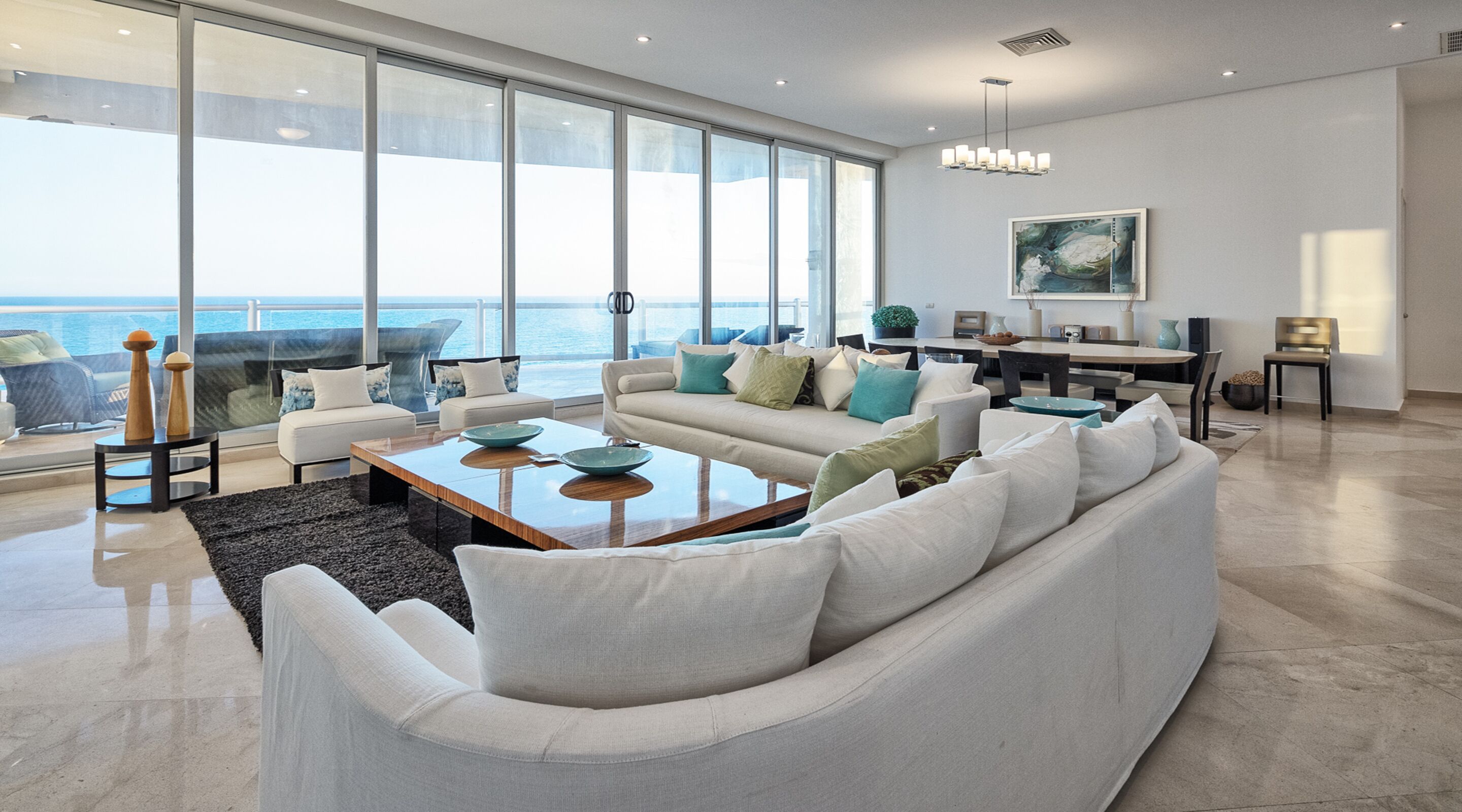 A spacious living room with a large window overlooking the ocean, a modern chandelier, and a dining area with a view.