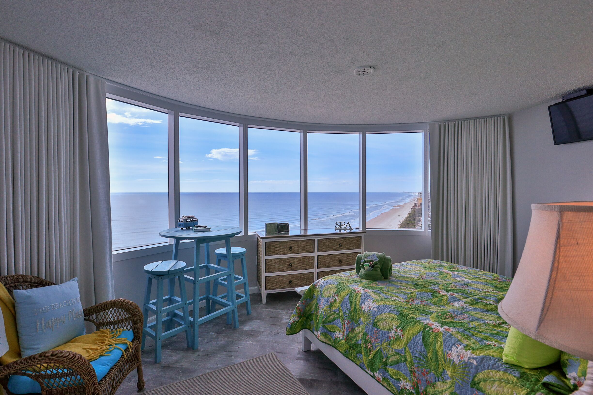 A bedroom with a curved window overlooking the beach, a bed with a floral bedspread, a small table with chairs, and a dresser.