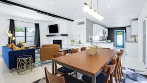 Un salon moderne avec un canapé bleu, une table à manger en bois et des chaises. Un téléviseur à écran plat est fixé au mur, il y a une cheminée et on aperçoit un coin cuisine à travers une porte ouverte.