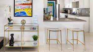 Une cuisine moderne avec des meubles blancs, un îlot central en acier inoxydable et une table basse en verre transparent aux pieds dorés.
