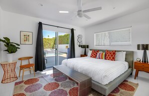 Une chambre moderne avec un grand lit, un ventilateur de plafond, une porte-fenêtre coulissante donnant sur un balcon et une vue sur la verdure environnante.