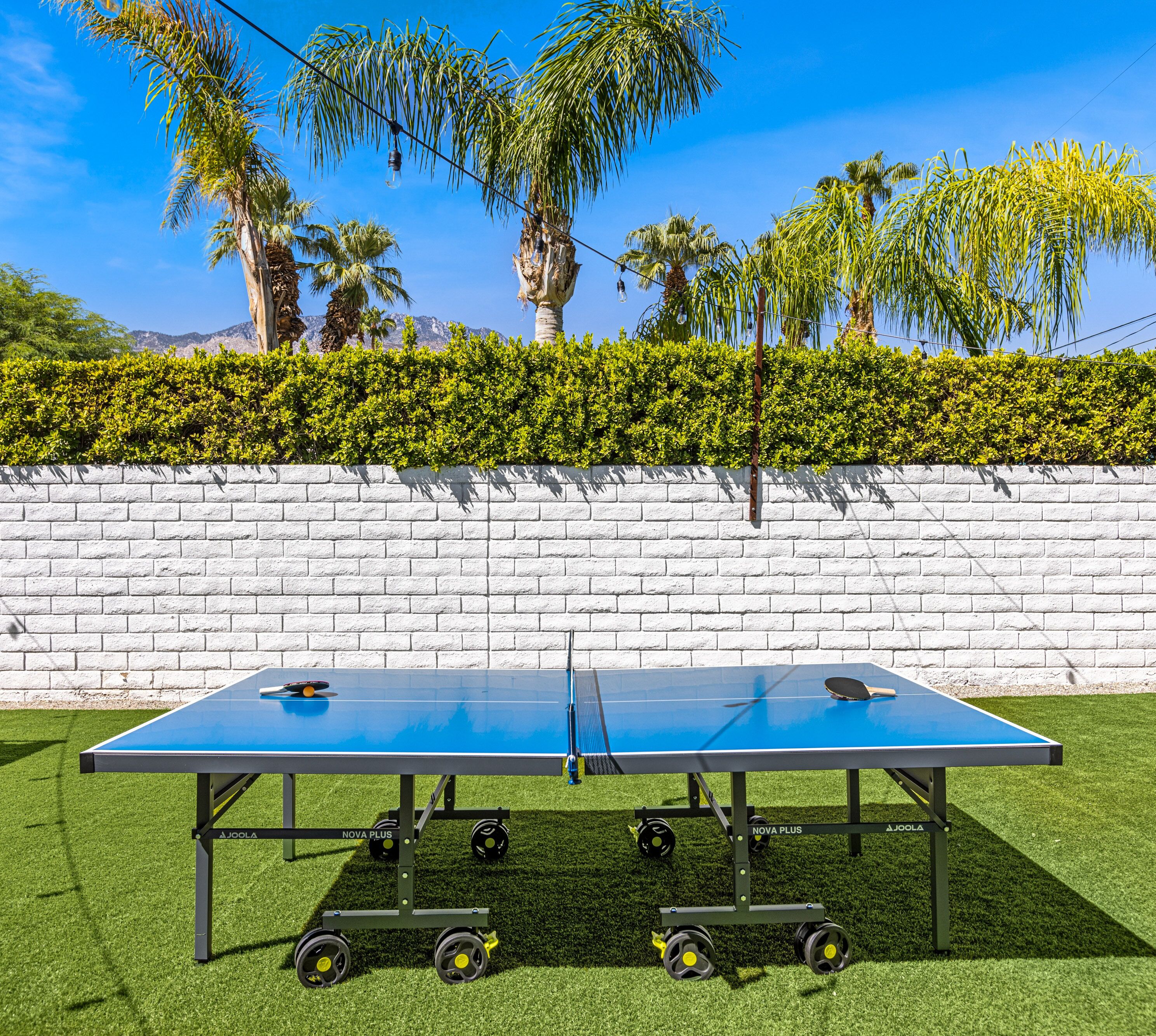 Una mesa de ping pong portátil con ruedas, montada sobre césped artificial, frente a una pared de ladrillo blanco con un seto y palmeras al fondo.