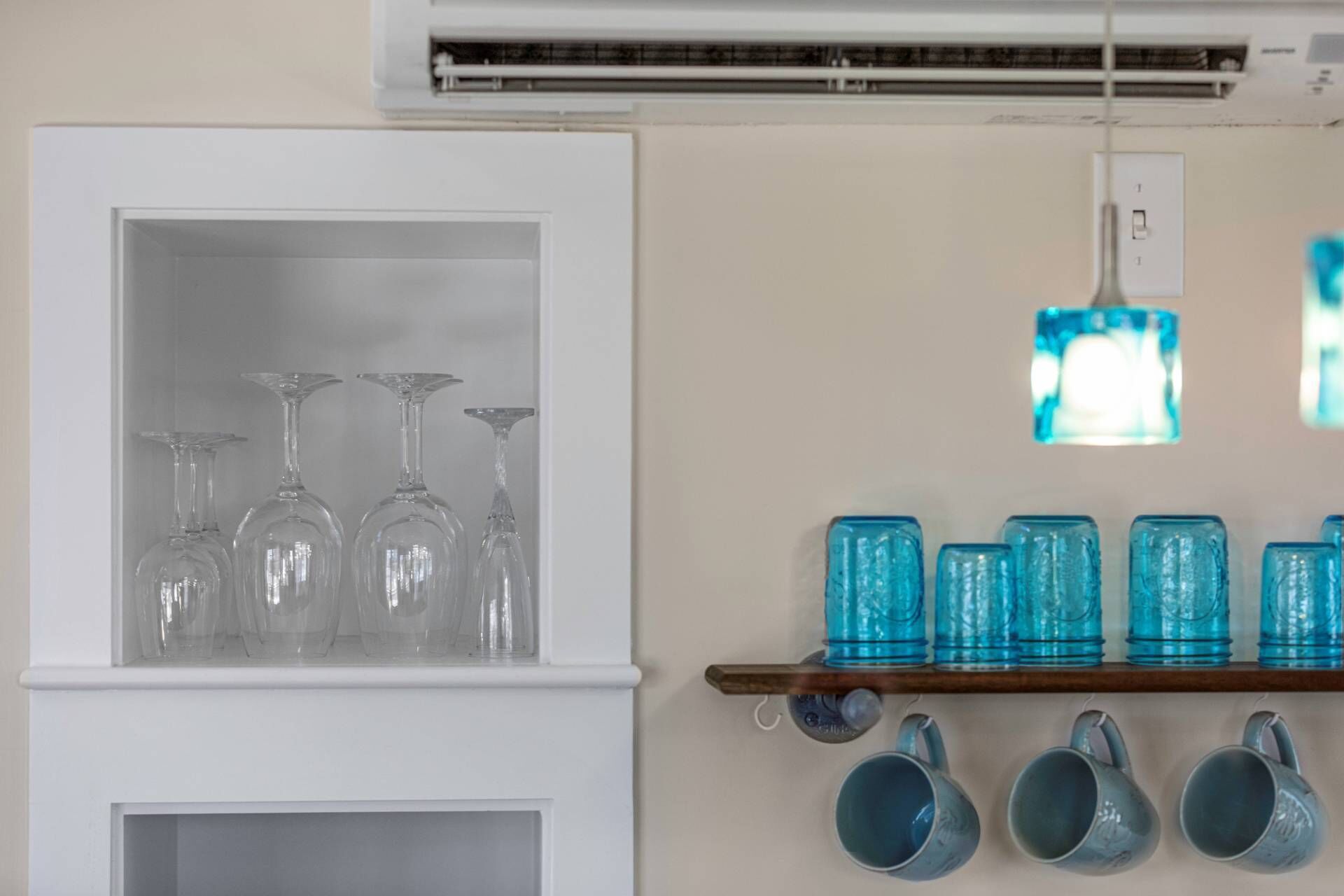 A kitchen with a white cabinet containing clear glassware and a shelf with blue glass mugs and a hanging light fixture.