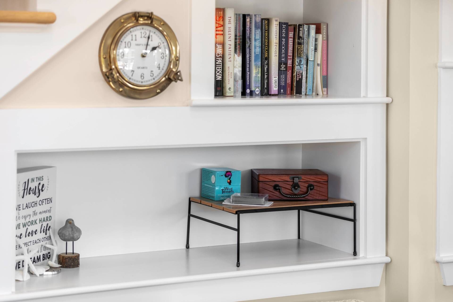 A bookshelf with books, a clock, a word teaser box, a bird figurine, and a wooden box on a white shelf.