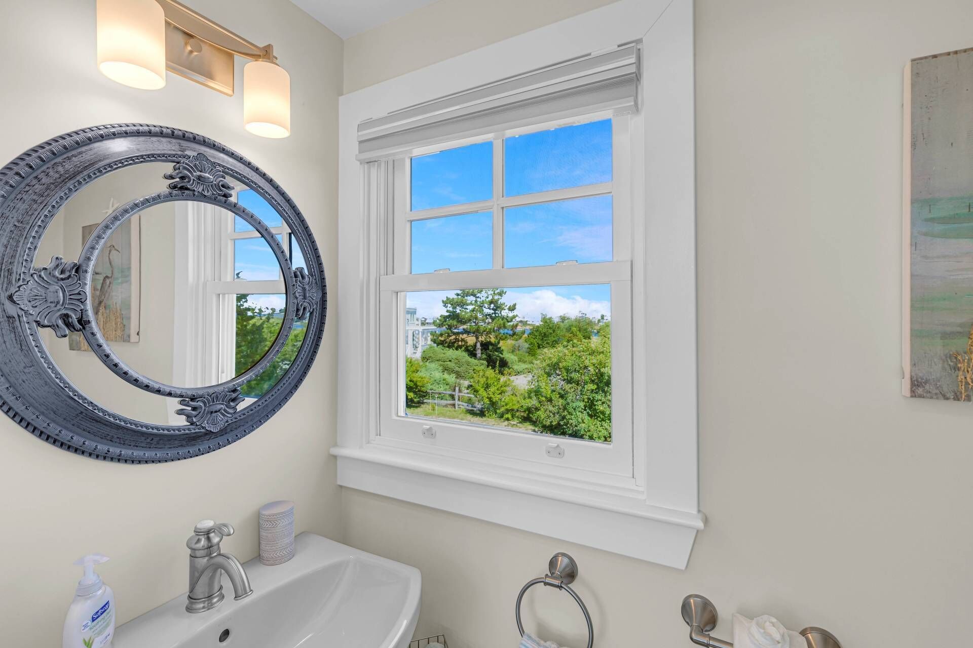 A bathroom with a large ornate mirror, a white sink, a window with a view of greenery, and a towel rack.