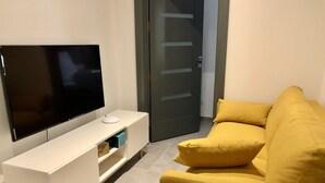 Een woonkamer met een flatscreen televisie op een witte dressoir, een gele bank met kussens, en een gesloten deur op de achtergrond.