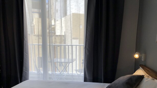 Hotelkamer met een bed, een raam met dunne gordijnen, een balkon met een leuning en een wandlamp.
