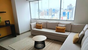 A modern living room with a sofa, a coffee table, and a view of a cityscape through large windows.