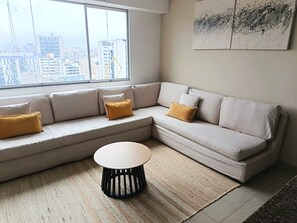 Un salon moderne avec un canapé d’angle, une table basse ronde et une grande fenêtre donnant sur la ville.