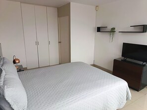 A bedroom with a bed, a wardrobe, a television on a stand, and a small table with a lamp.
