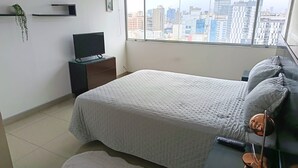 A bedroom with a bed, a TV, a nightstand, a window with a city view, and a small plant.