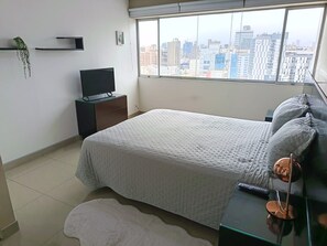 Une chambre à coucher avec un lit, une télévision, une table de chevet, une fenêtre donnant sur la ville et une petite plante.