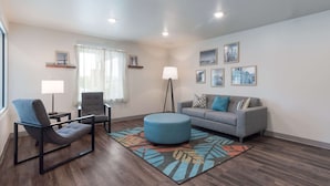 A modern living room with a grey sofa, blue armchair, and ottoman, a floor lamp, and framed pictures on the walls. - Woodspring Suites Philadelphia Northeast (Philadelphia)