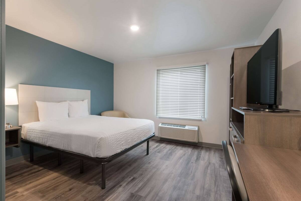 A modern hotel room with a bed, bedside table, lamp, air conditioning unit, window with blinds, and a TV on a wooden cabinet.