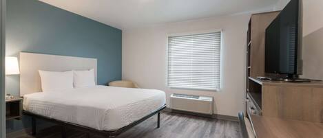 A modern hotel room with a bed, bedside table, lamp, air conditioning unit, window with blinds, and a TV on a wooden cabinet.