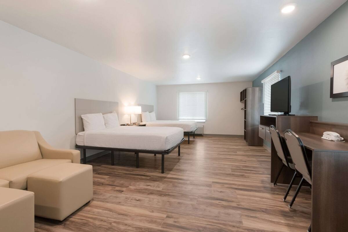 A modern hotel room with a large bed, two smaller beds, a sofa, a desk with chairs, a TV, and a window with blinds.