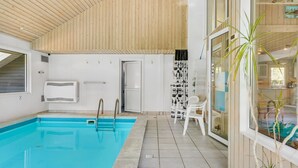Une piscine intérieure moderne avec un plafond en bois, une porte vitrée donnant sur une pièce et une vue sur l’extérieur.