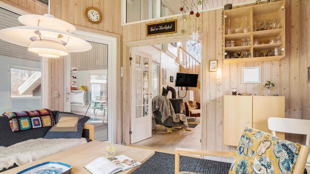 Une pièce chaleureuse avec un décor en bois, un canapé, un fauteuil, une table avec un livre et un verre, et une pancarte sur laquelle on peut lire : « Gud ou Karlshad ».