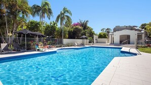 Een zwembad met helderblauw water, omringd door ligbedden en parasols, palmbomen en een modern gebouw op de achtergrond.
