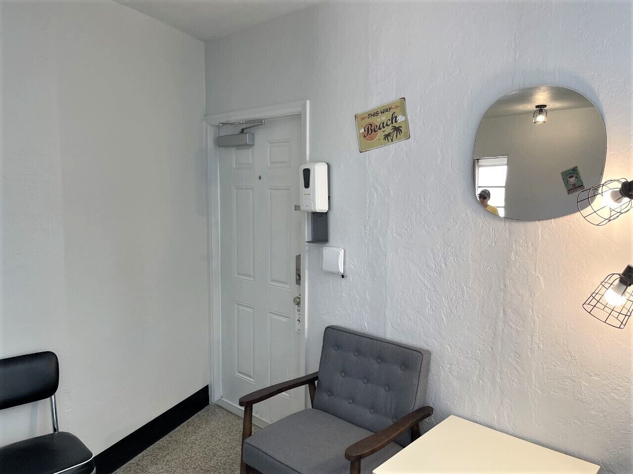 Ein Zimmer mit einer weißen Tür, einem grau gepolsterten Sessel, einem kleinen Tisch und einem wandmontierten Spiegel.