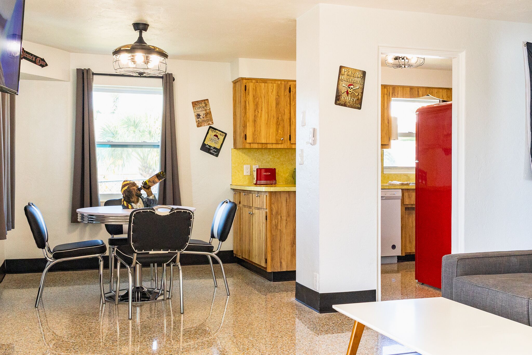 A dining area with a round table and four chairs, a kitchen with wooden cabinets and a red refrigerator, and a living area with a grey sofa.