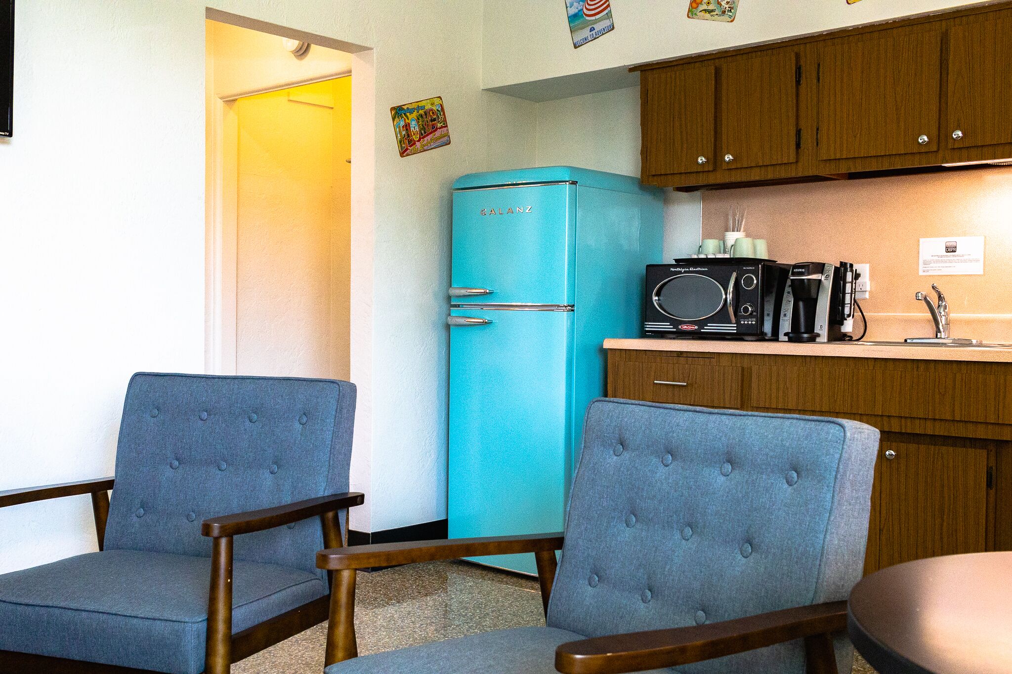 A compact kitchen with a turquoise refrigerator, wooden cabinets, and two blue upholstered chairs.