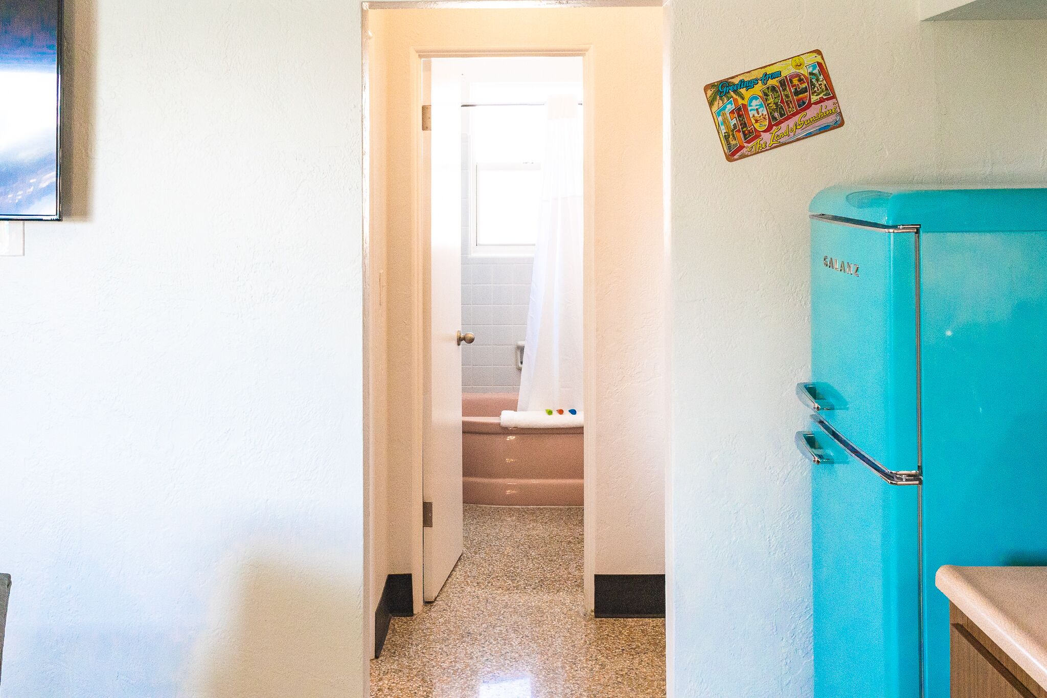 Un pasillo con la puerta de un baño que da a un inodoro y una bañera. Se ve un refrigerador azul a la derecha.