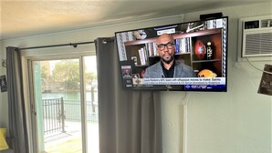 Een kamer met een aan de muur bevestigde televisie waarop een sportwedstrijd wordt vertoond. Er is een boekenkast met boeken en een voetbal. Een raam met gordijnen is zichtbaar, evenals een deur die naar buiten leidt.