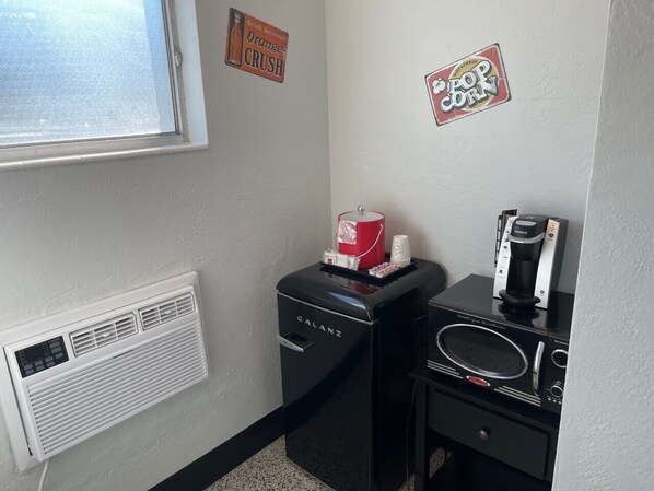 A small kitchenette with a coffee maker, a kettle, and a microwave. There is a window with a view of the outside, and a wall-mounted air vent.