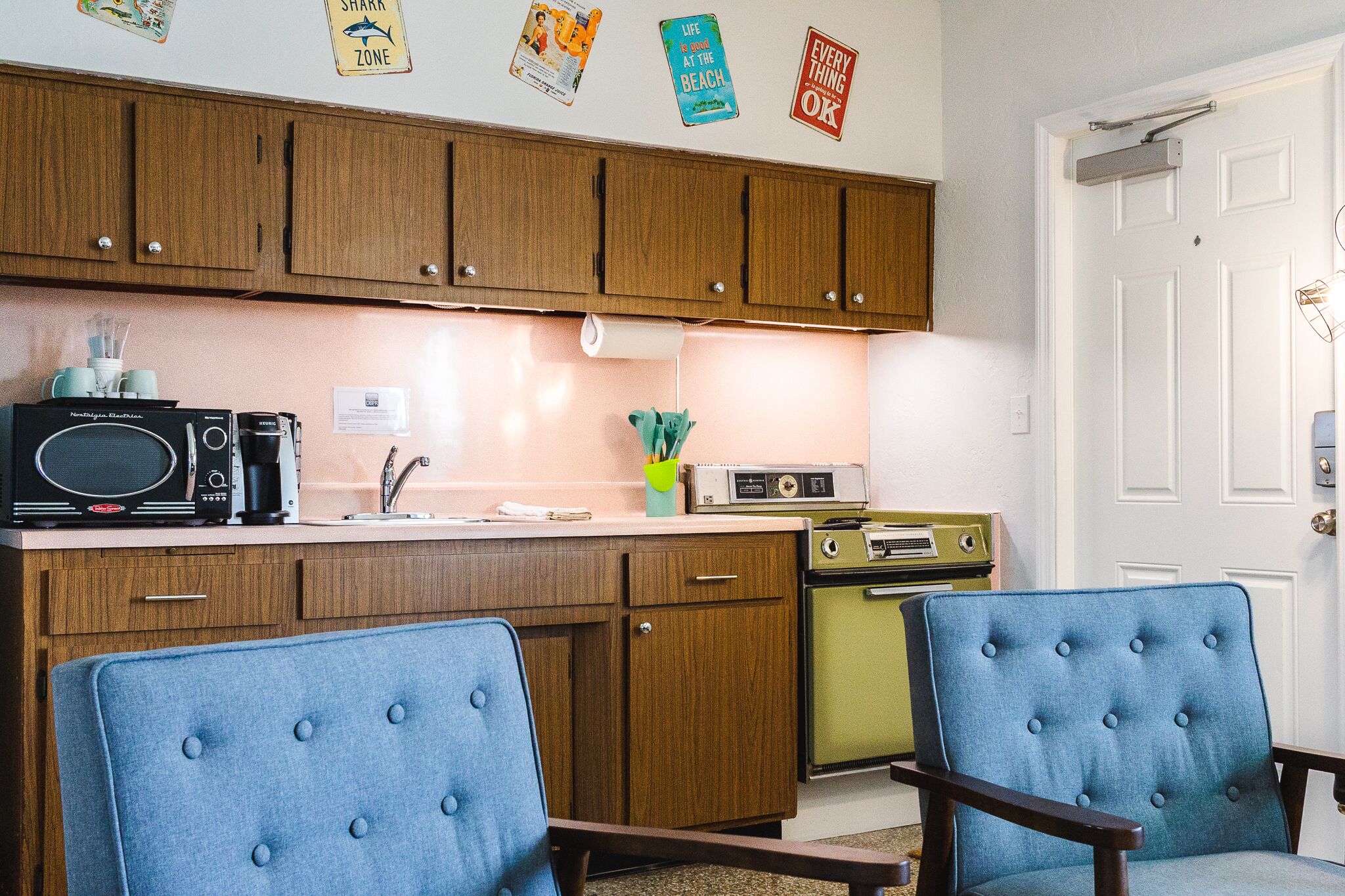 A compact kitchen with wooden cabinets, a microwave, a stove, and two blue upholstered chairs.