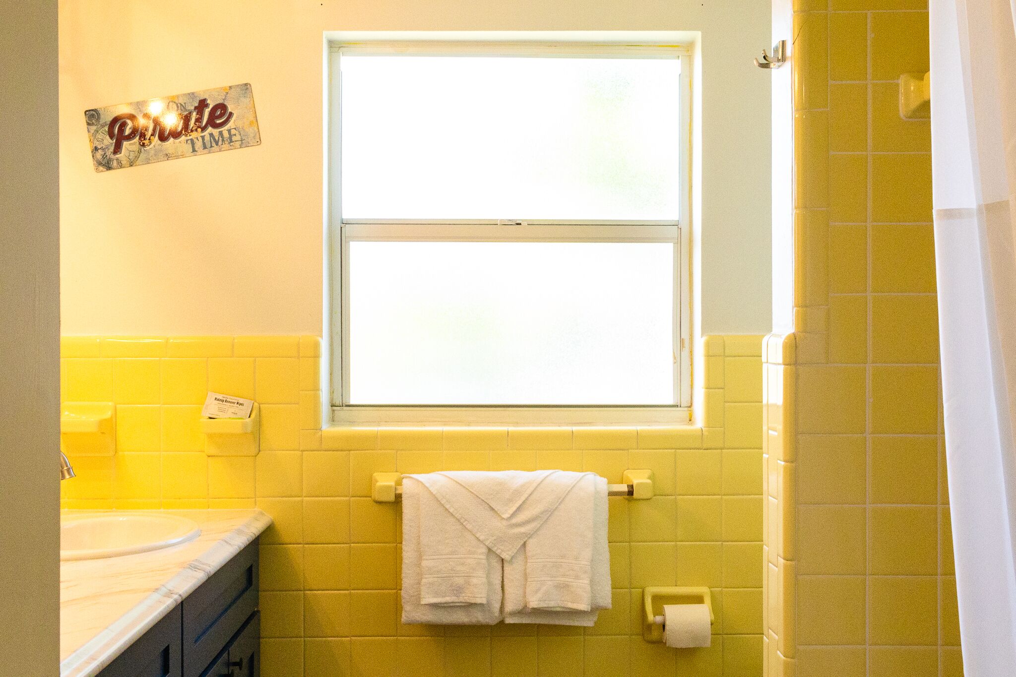 Een badkamer met gele tegels, een raam, een handdoekrek met een handdoek en een bordje aan de muur.