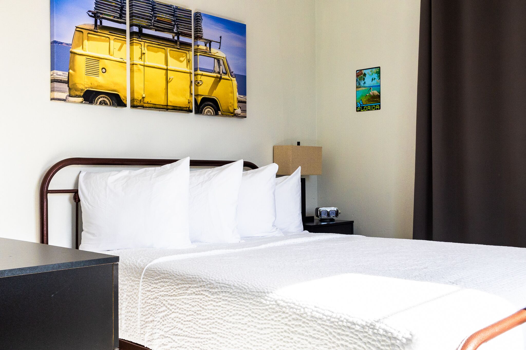 A hotel room with a bed, a nightstand, a lamp, and a wall with a van and Florida poster.