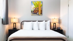 Een netjes opgemaakt bed met witte lakens, twee bedlampjes en een schilderij van een auto en een palmboom aan de muur.