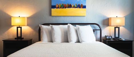 A hotel room with a bed, two bedside tables, a painting of people on a beach, and a nightstand with a clock.
